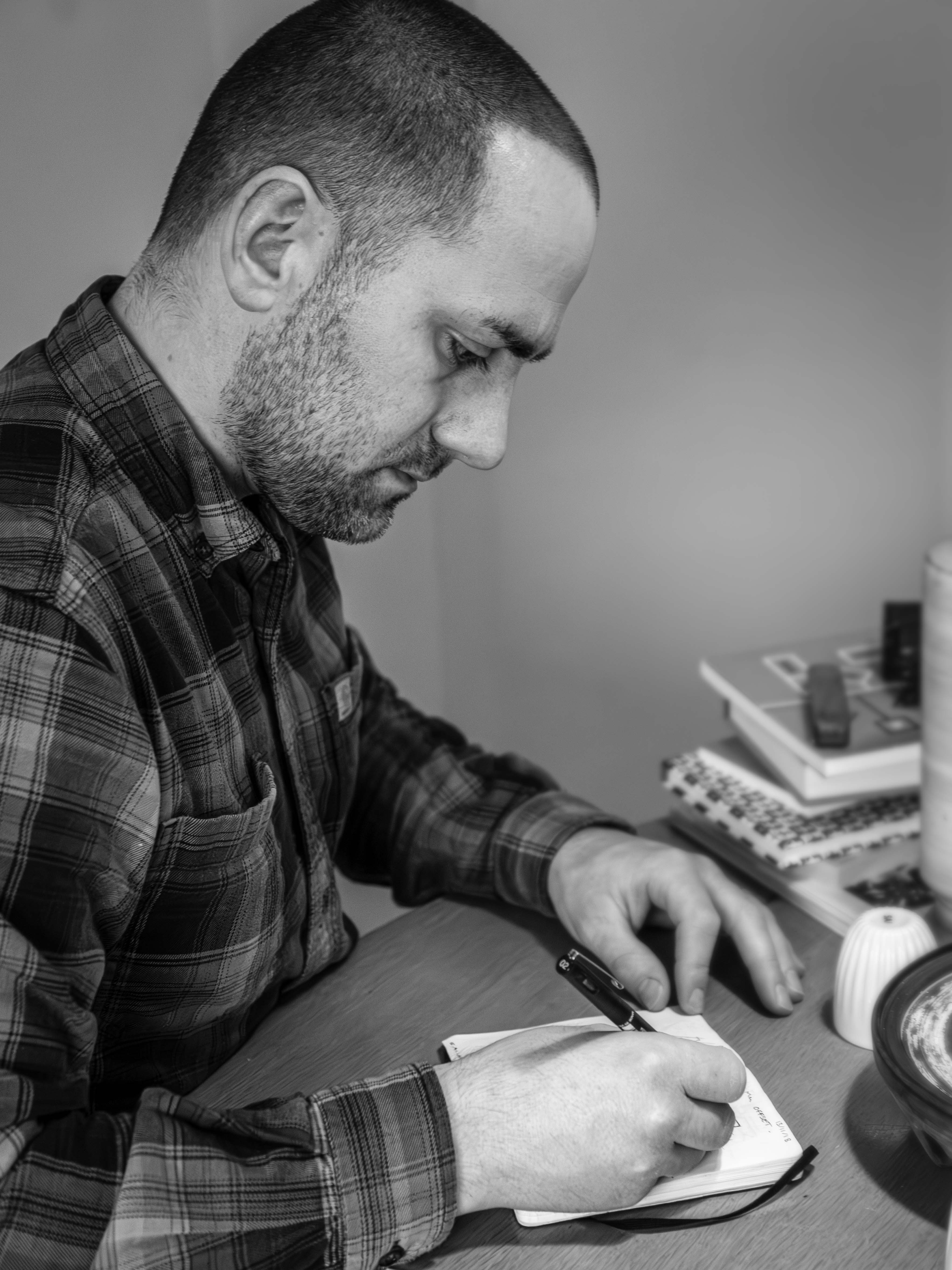 Man in plaid shirt writing on a piece of paper with books and a cup in the background